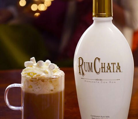 Une tasse en verre de chocolat chaud garnie de creme fouettee et une bouteille de rumchata