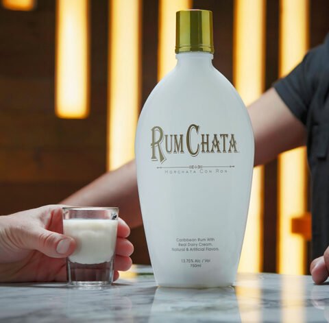 Someone holding a shot of chilly-chata with a bottle of Rumchata on a marble table