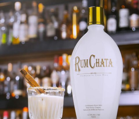 A shot of cinnamon toast with cinnamon stick in it and a bottle of Rumchata with a wall of spirit bottles background