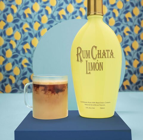 Une tasse en verre de latte chaud au limon et une bouteille de rumchata limon devant un papier peint de citron et feuilles blueues