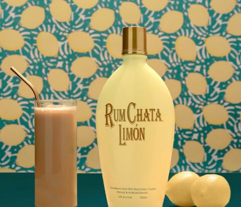 A glass of iced lemon latte with a bottle of Rumchata Limon and two lemon in front of a lemon wallpaper