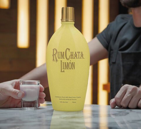 Someone holding a shot of black limon berry on a marble bar table with a bottle of Rumchata Limon