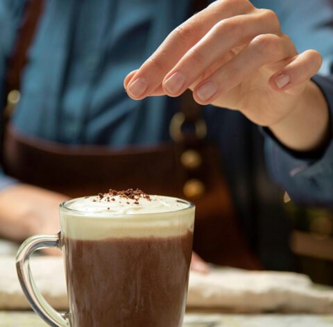 Someone adding a pinch of grated chocolate on top of a spiced mexican coffee in a glass cup